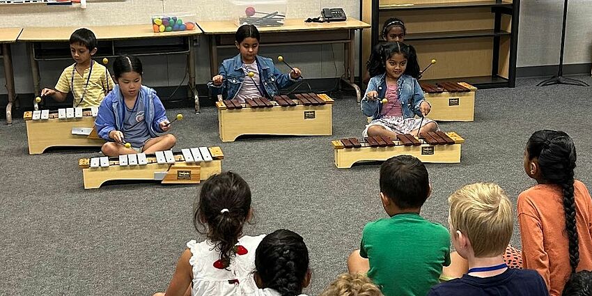 4 young students sitting on the floor playing xylophones while their classmates watch and listen