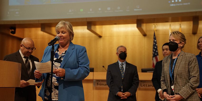 woman at podium. People standing behind her listening.